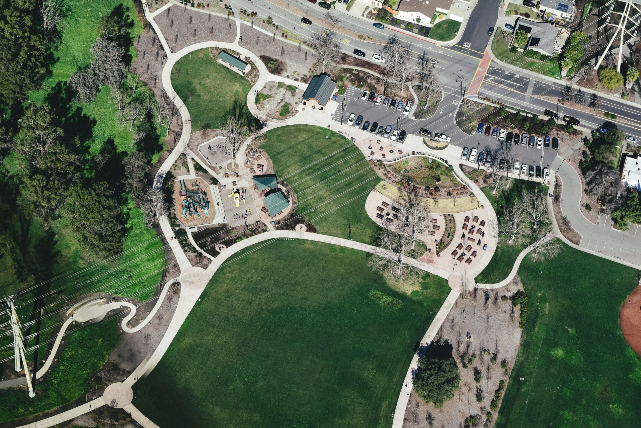 Drone view of modern city park area with winding roads surrounded by greenery and cars parked nearby located under power lines