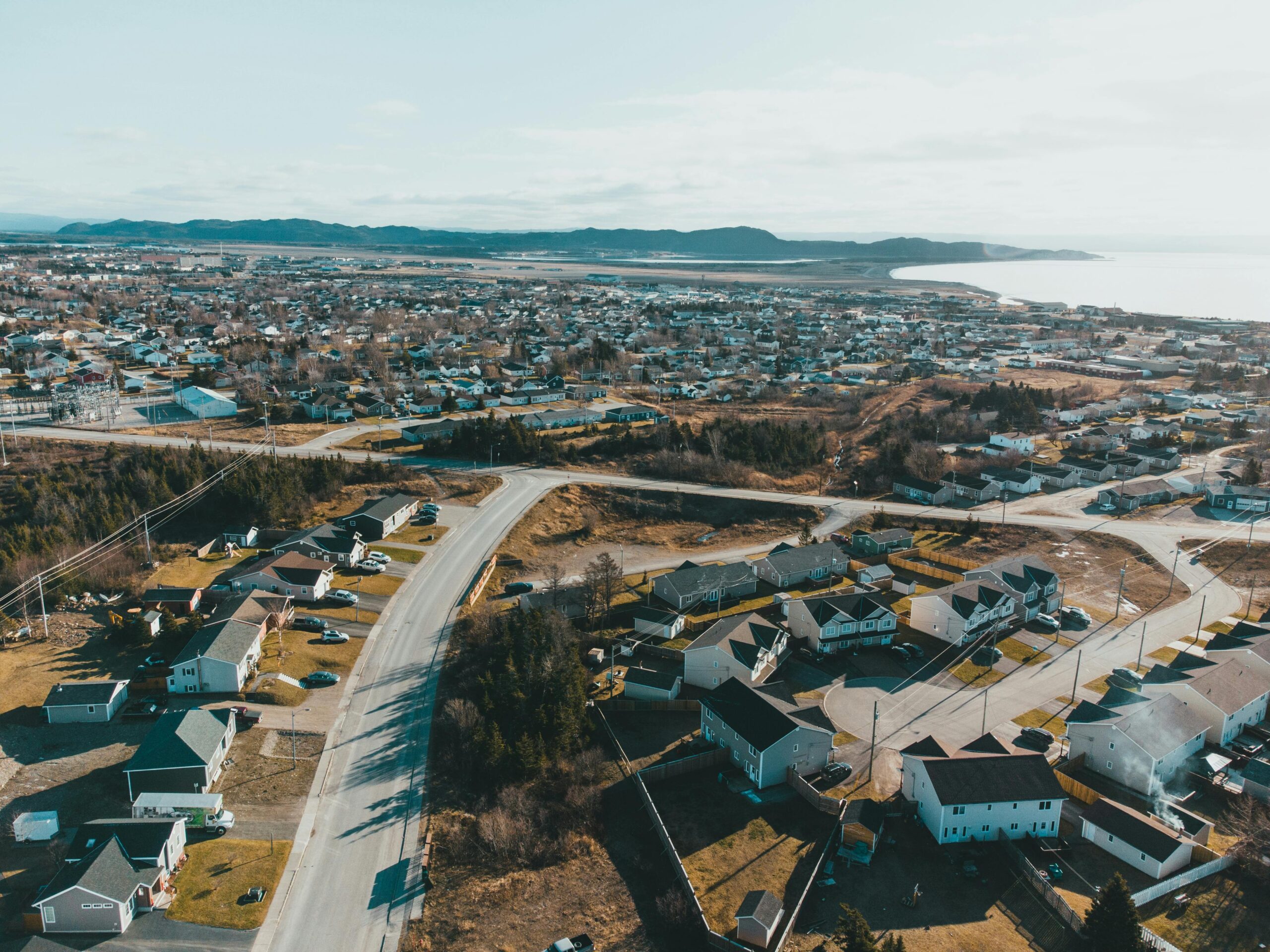 Drone view of coastal settlement with small houses and asphalt road near sea and mountain on background under light blue sky on sunny day in autumn